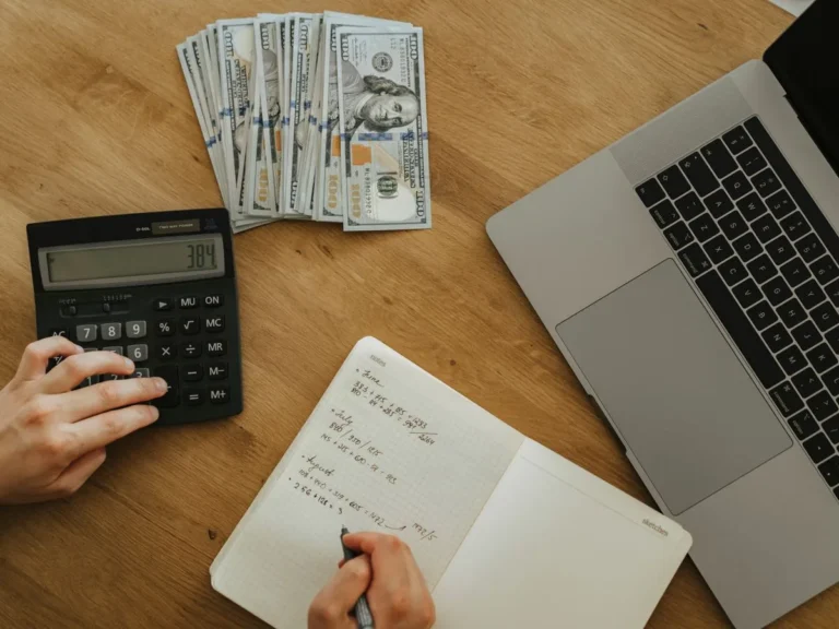 Manos usando una calculadora y anotando cifras en un cuaderno junto a un laptop, calculando el costo de un proyecto